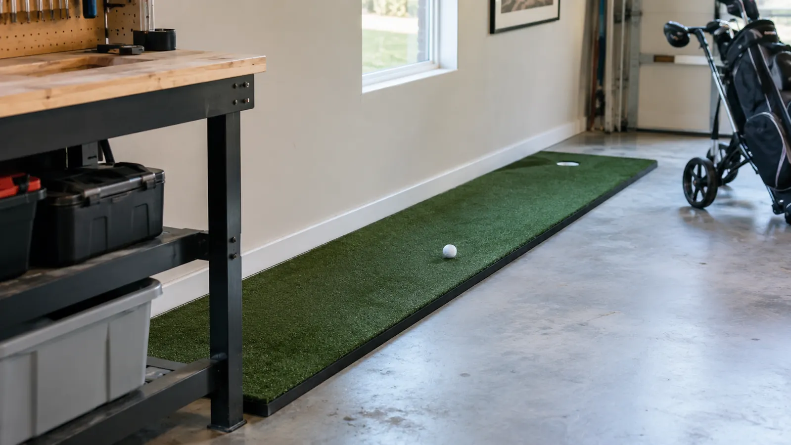 A 3 foot by 9 foot indoor putting green rolled out flat along a garage wall, a golf ball resting near the cup