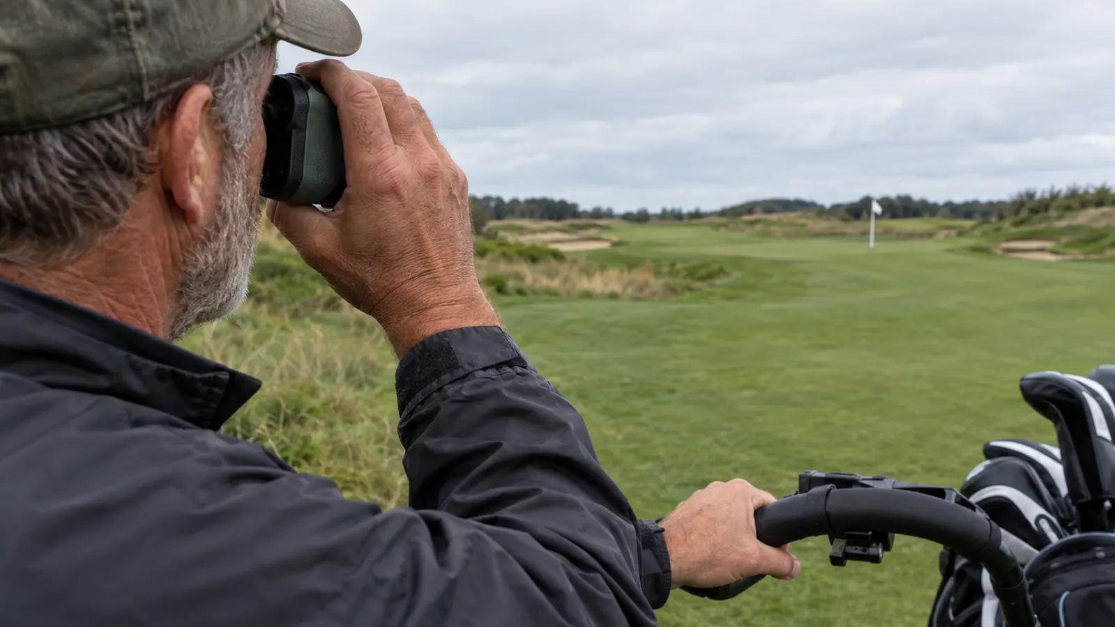 A golfer's hand holding a rangefinder steady against a push cart handle while aiming at a flagstick