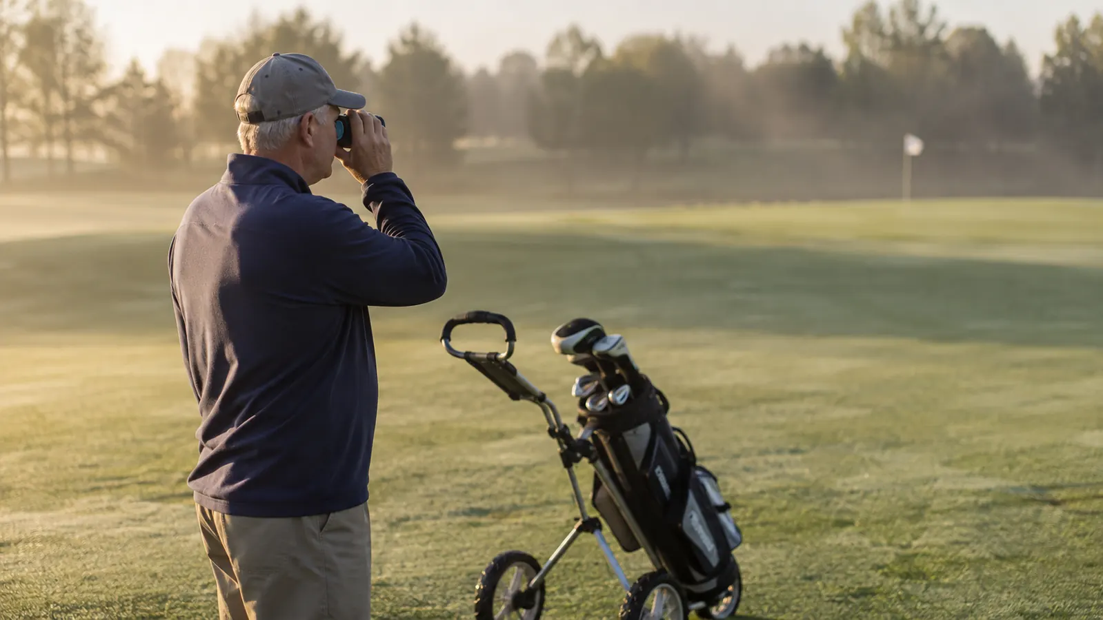 A golfer on an early morning fairway checking the distance to the green through a laser rangefinder, push cart beside him